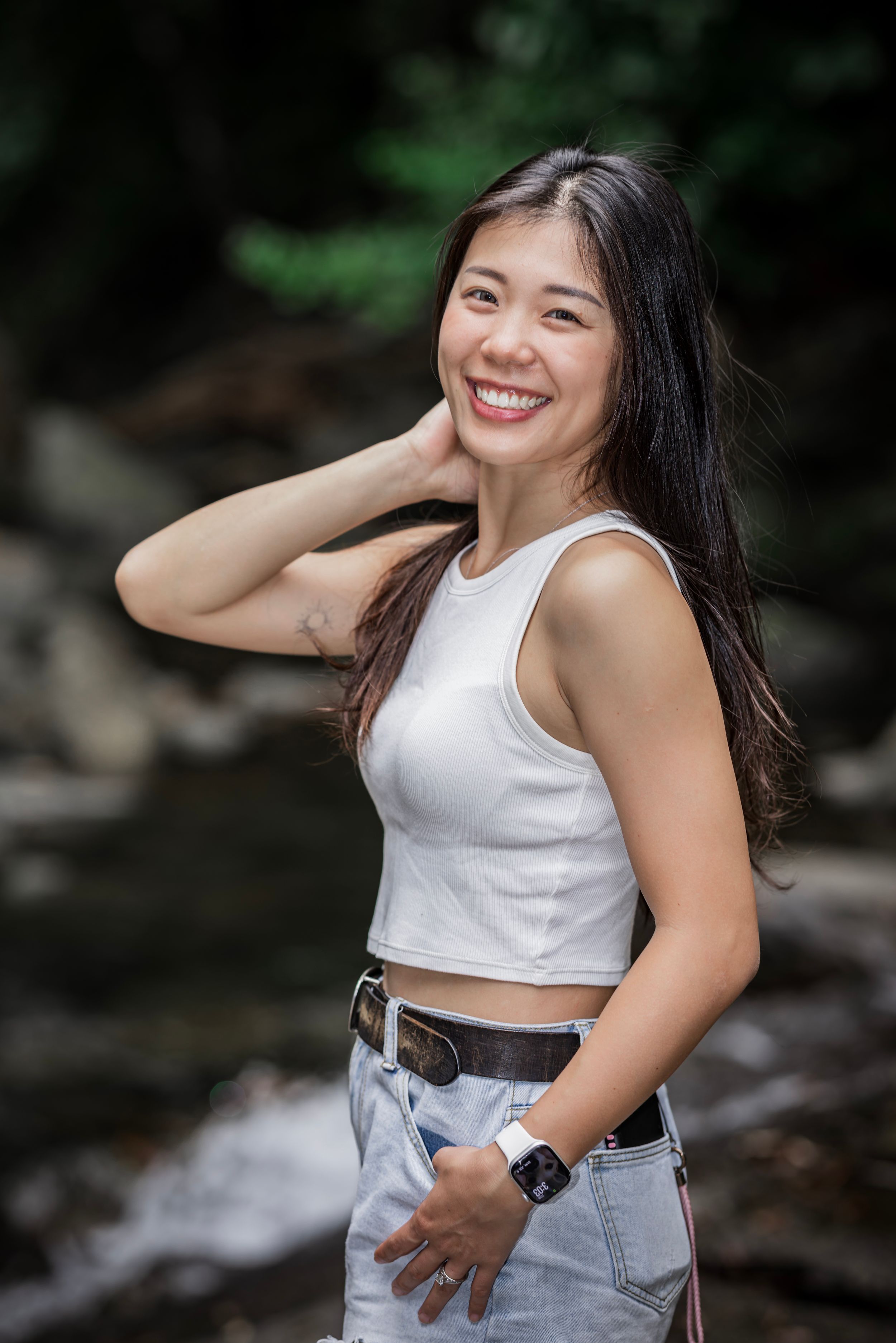 A woman in a white crop top and jeans poses outdoors near a stream with a bright smile and natural backdrop.