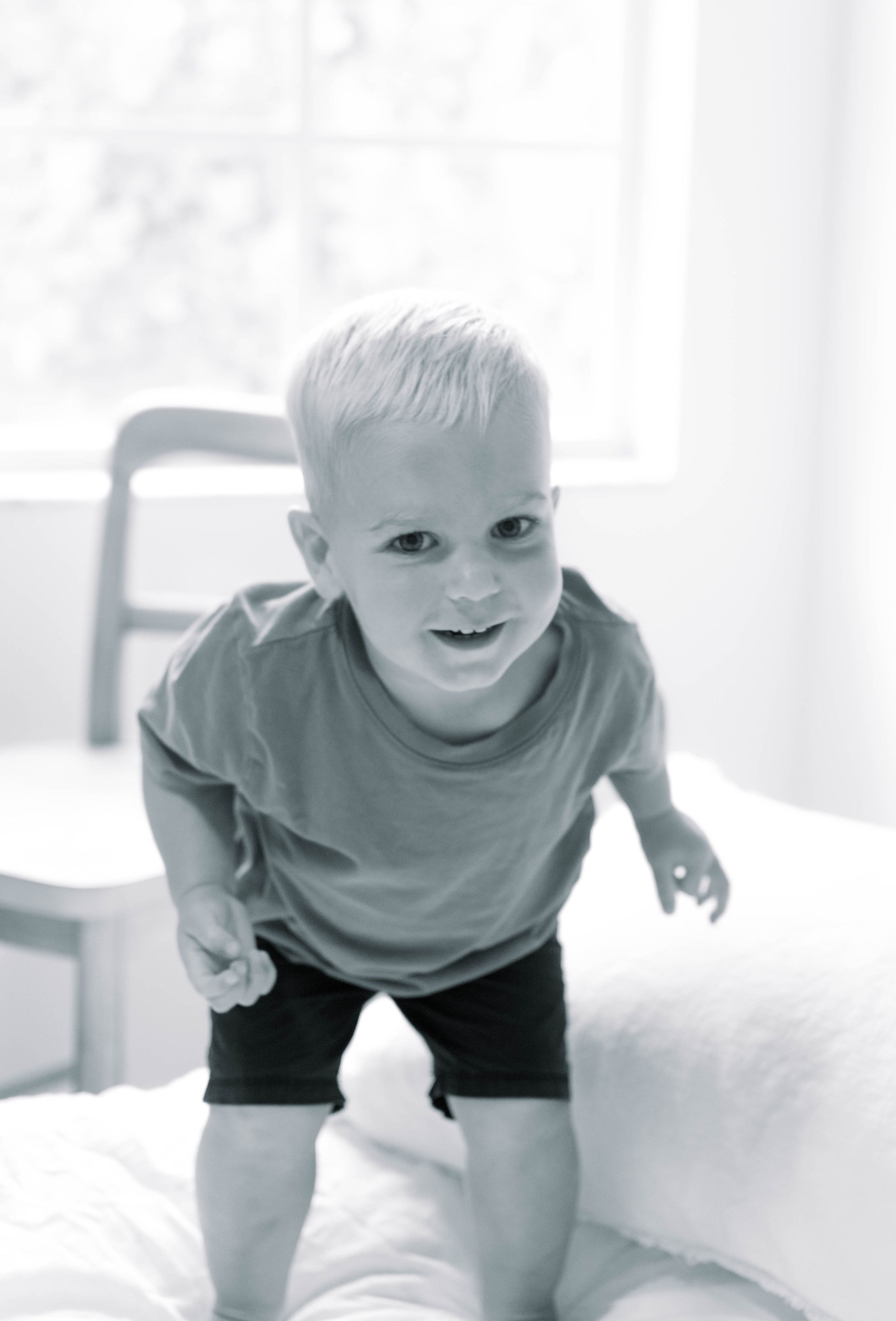 Black and white photo of a child in a t-shirt and shorts smiling at the camera in a bright indoor setting.