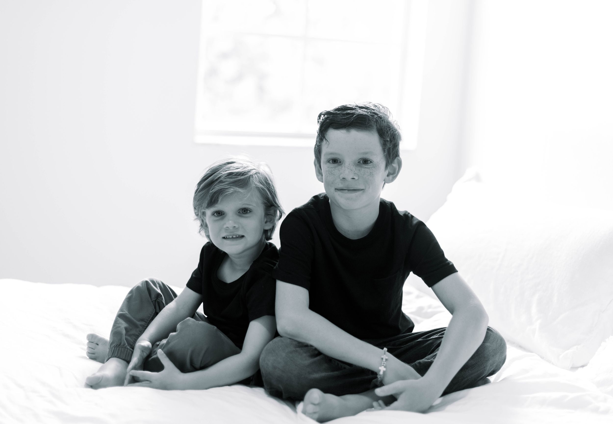 A black and white photo series of two casual boys relaxing together in white bedding.