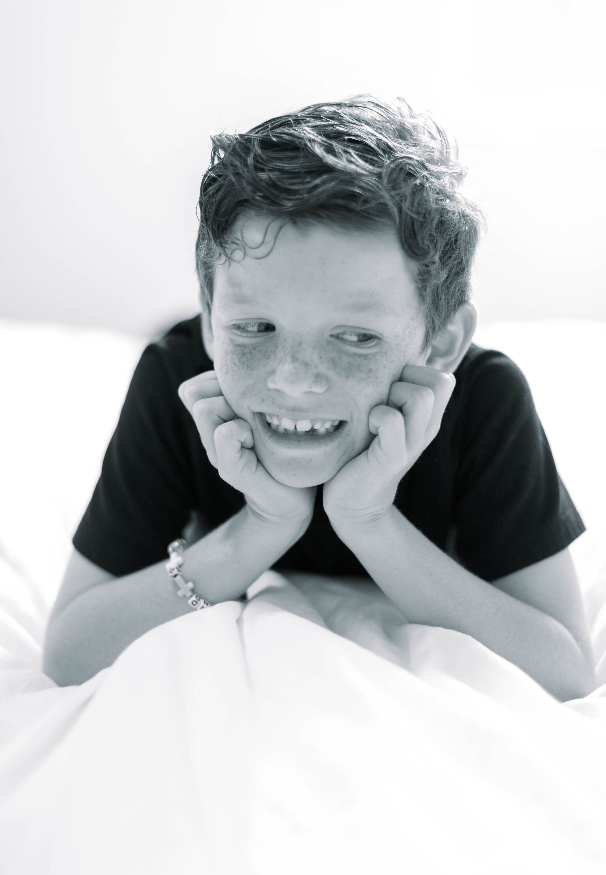 Black and white portrait of child lying on white bedding with hands under chin smiling at camera.