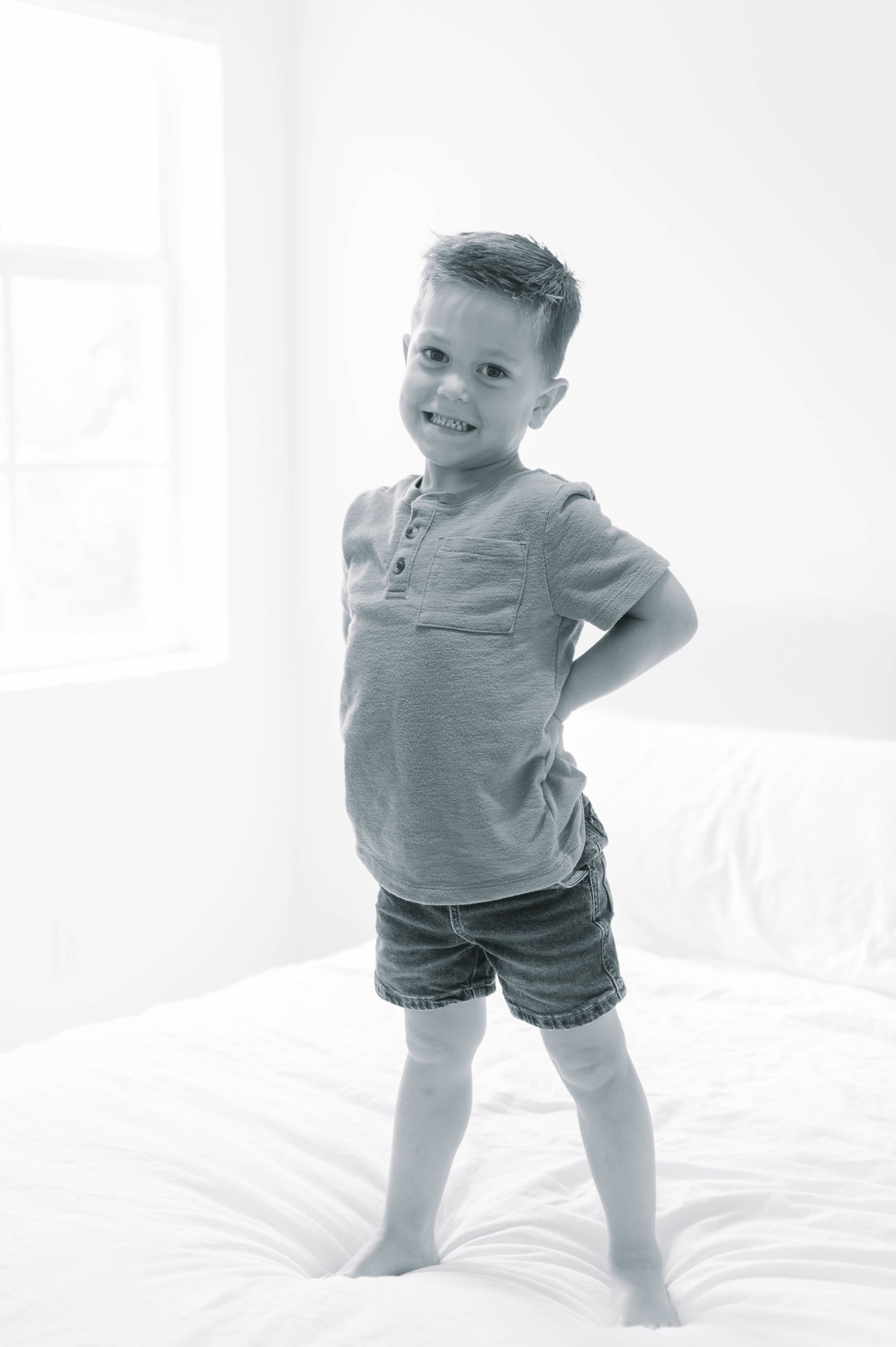 Black and white portrait of a young child standing on a bed wearing casual summer clothing.