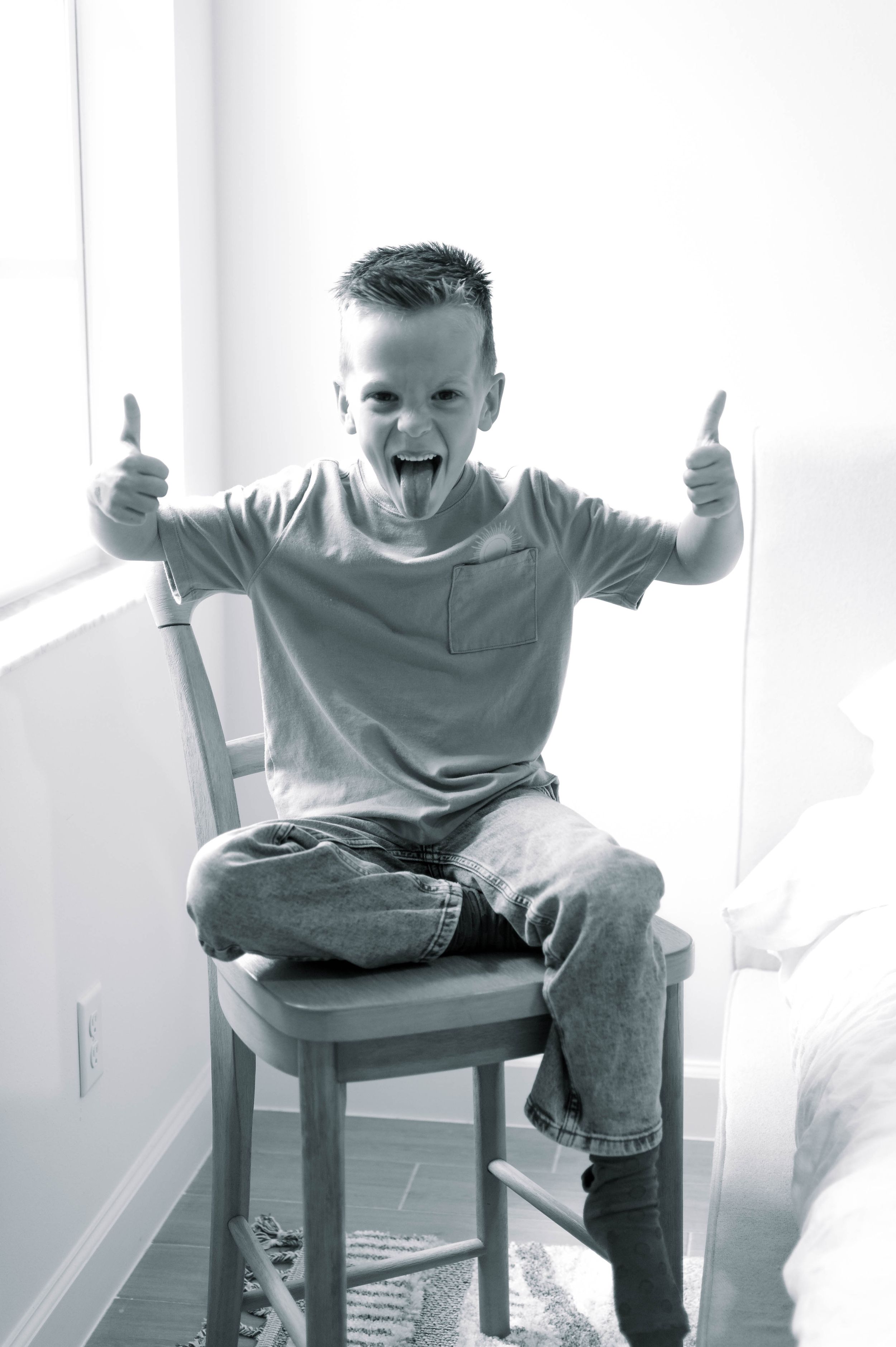 Black and white photo of a young child sitting on a wooden chair giving enthusiastic thumbs up gestures.