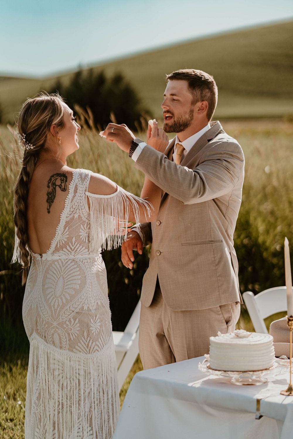 Bride and groom sharing a joyful cake tasting moment at outdoor boho wedding reception.