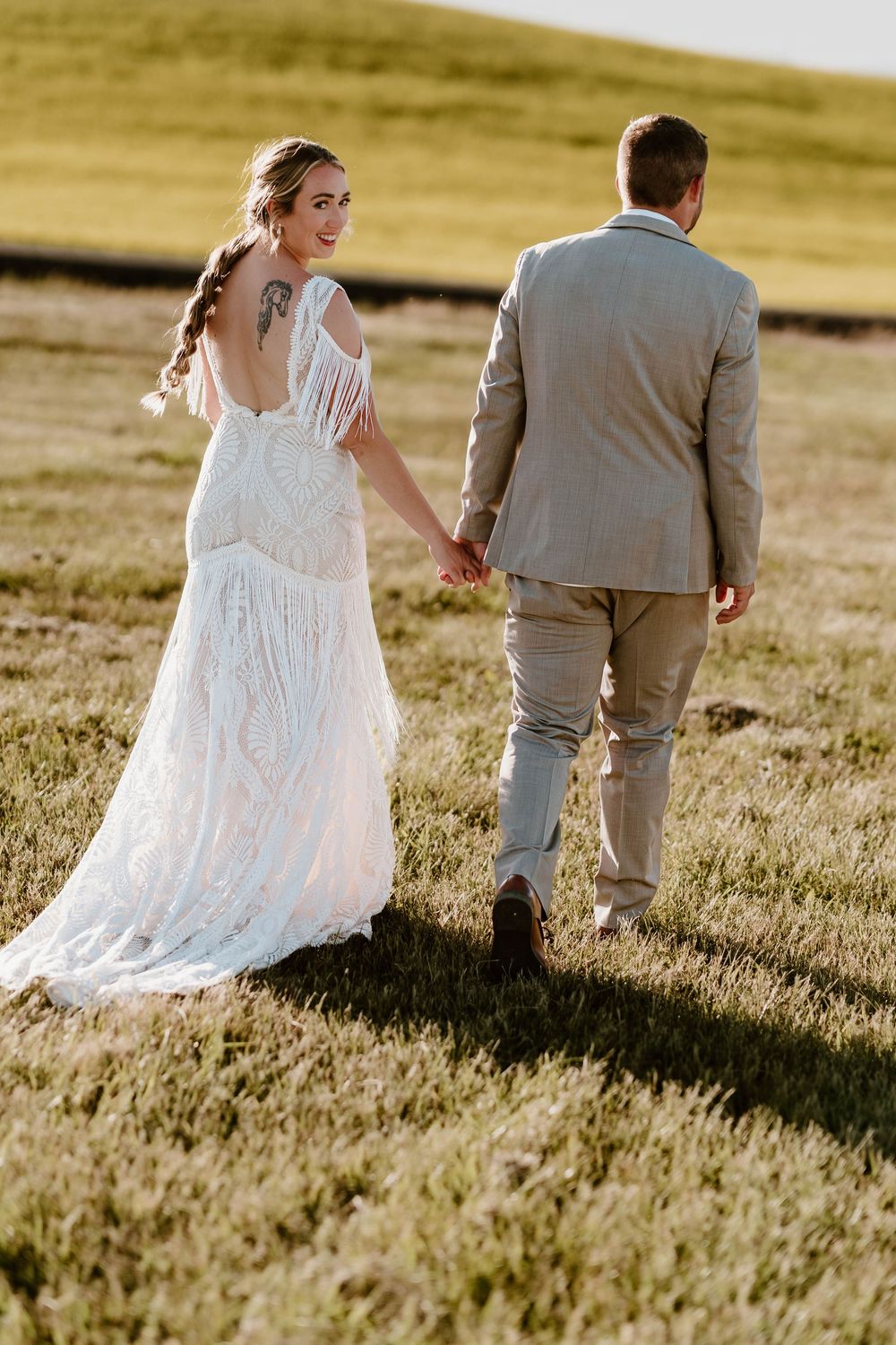 Bride in boho lace fringe gown holds hands with groom in tan suit walking through green field at golden hour.