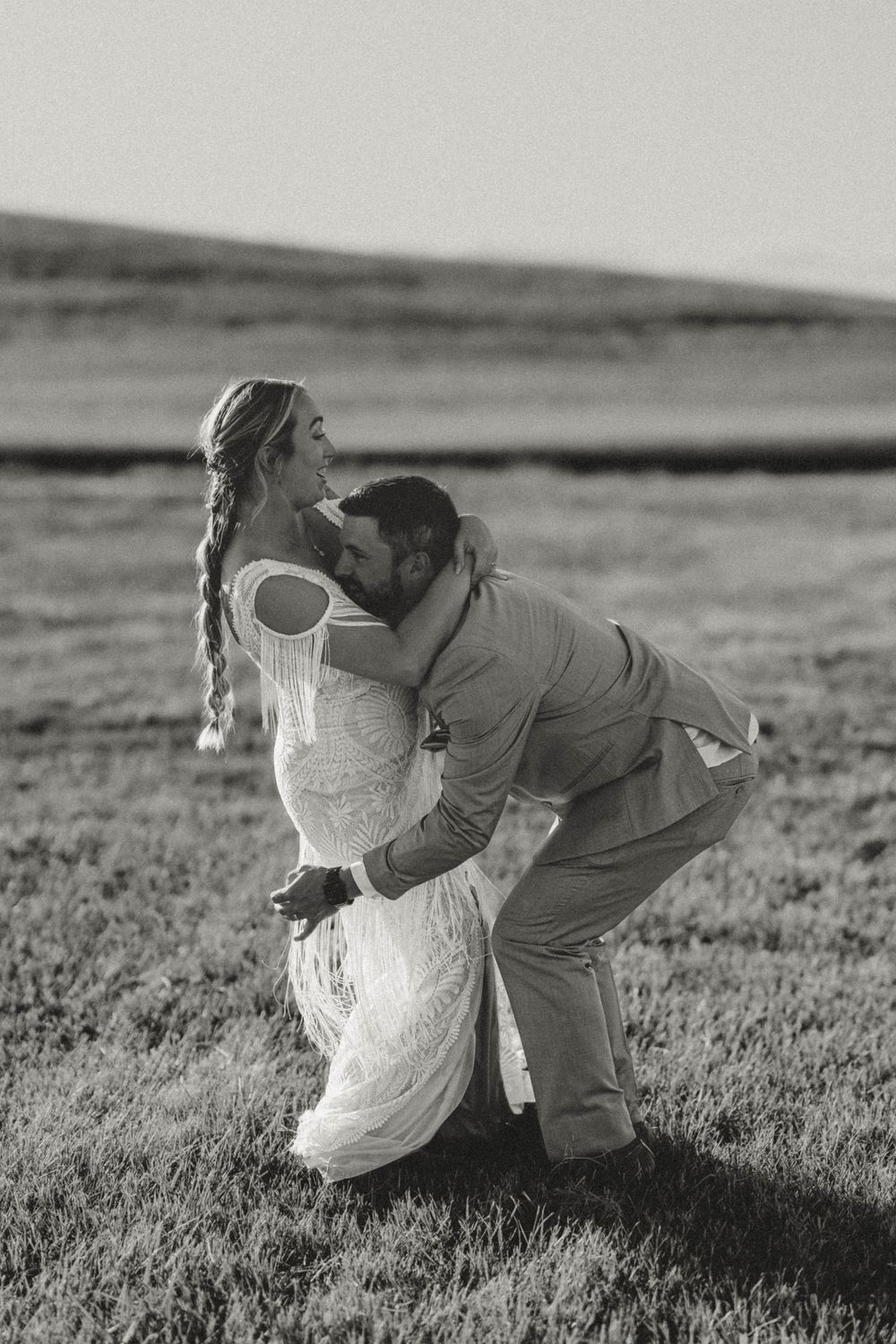 Black and white photo of groom lifting bride in boho wedding dress in an open grassy field.