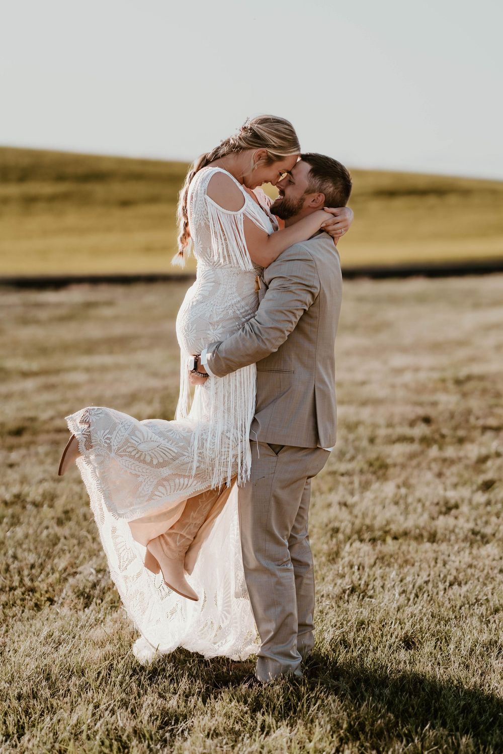 Groom lifts bride in boho lace fringe dress for a kiss in a sunlit open field at golden hour.