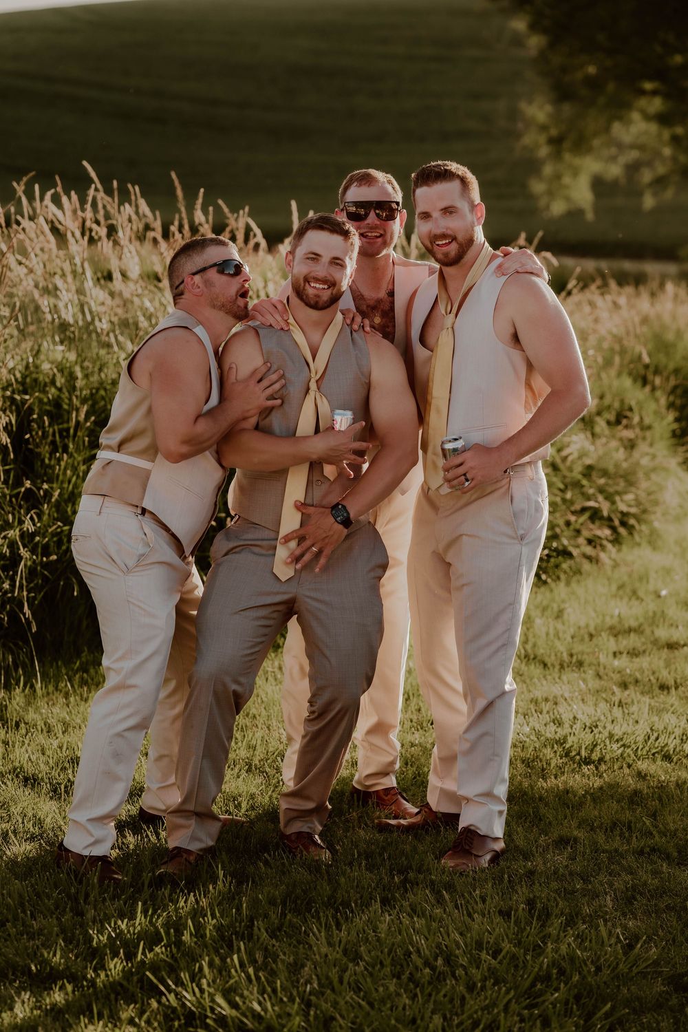 Four groomsmen in tan suits laughing and posing together outdoors at a summer wedding celebration.