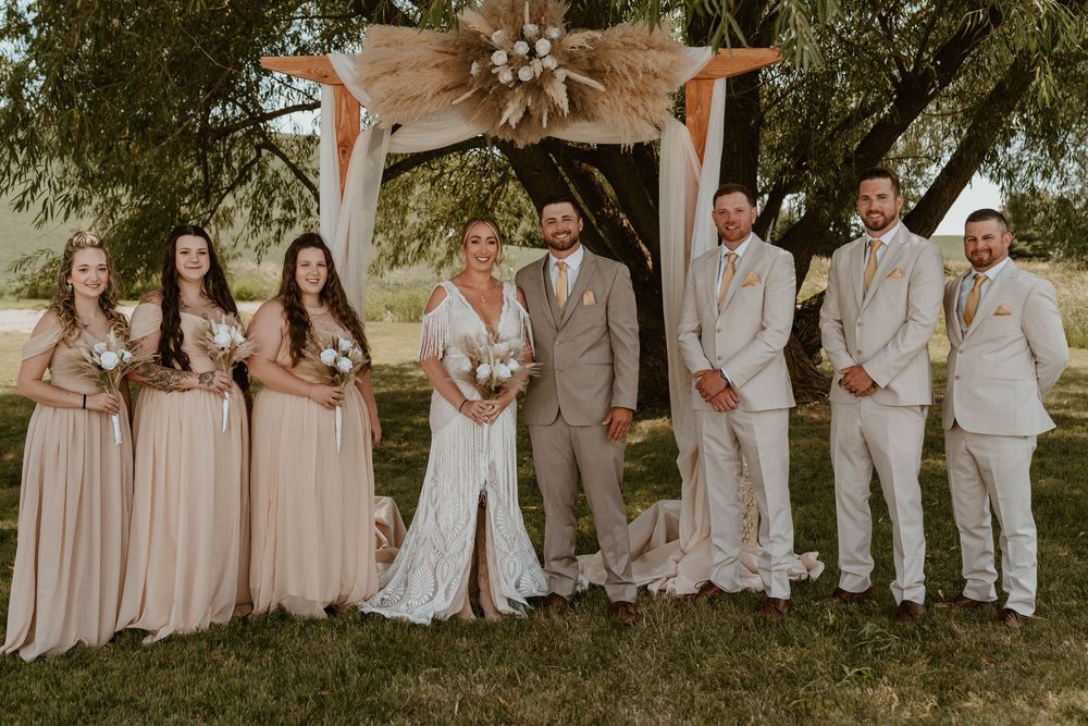 Wedding party poses under boho pampas grass arch with bridesmaids in blush gowns and groomsmen in tan suits.