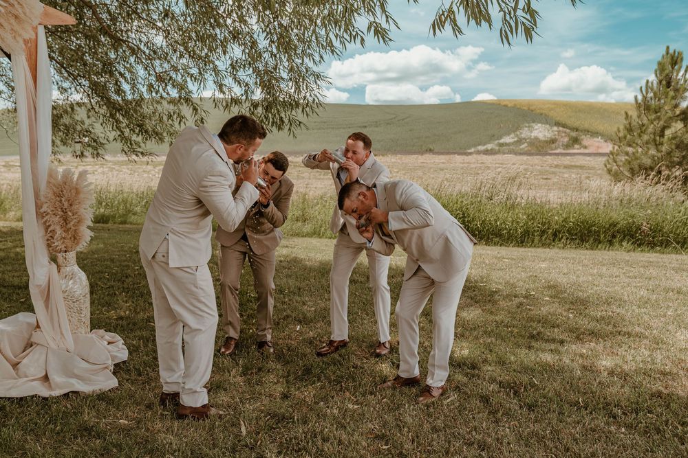 Groomsmen in tan suits playfully petting a dog on the grass near a boho wedding ceremony arch.