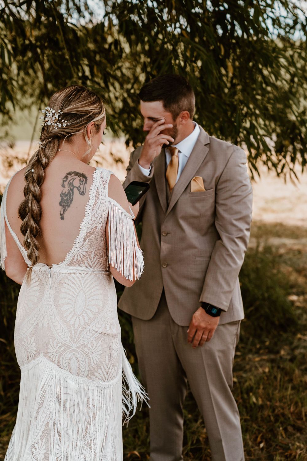 Emotional groom wipes tears seeing bride in boho fringe lace gown during their first look outdoors.