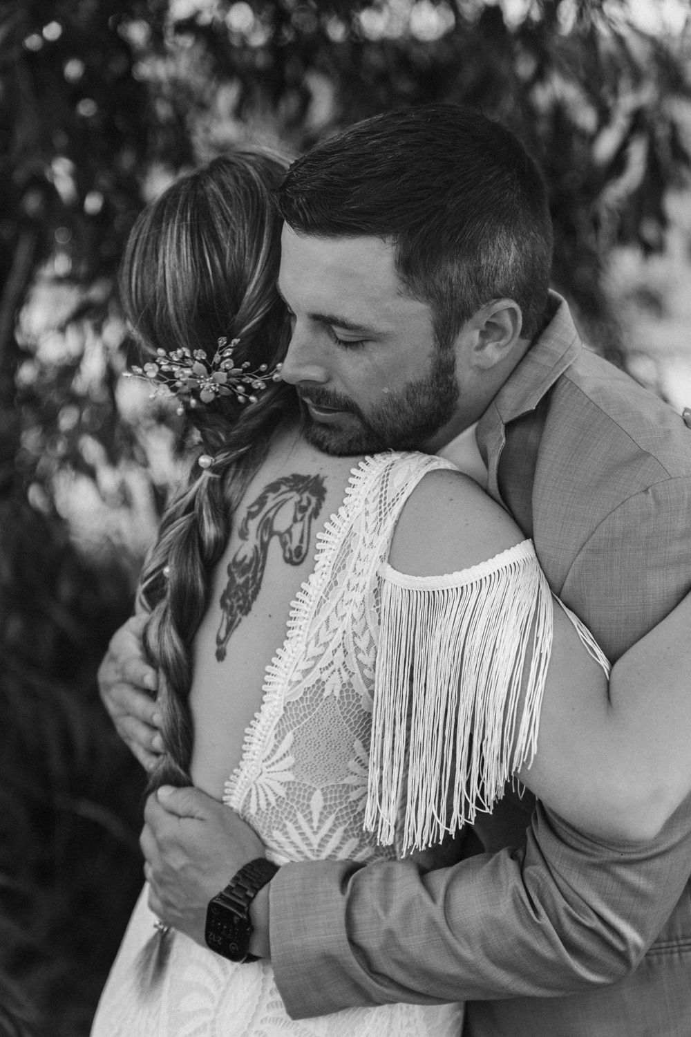Black and white photo of groom tenderly embracing bride in boho lace fringe dress under leafy trees.