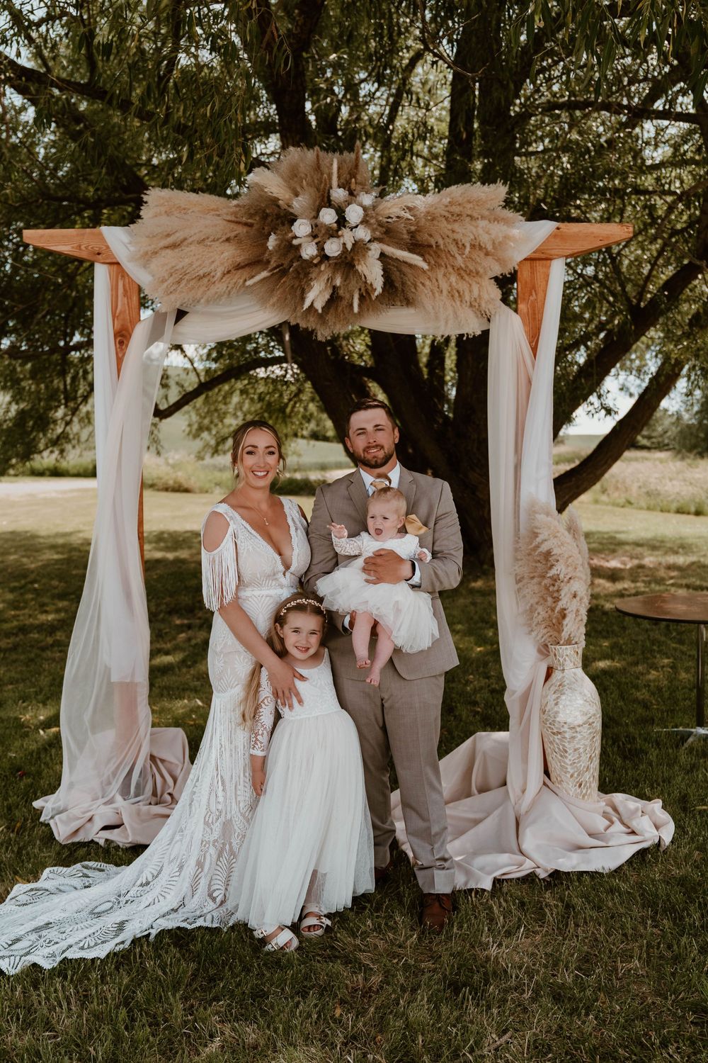 Bride and groom pose with two young children under a boho pampas grass wedding arch outdoors.