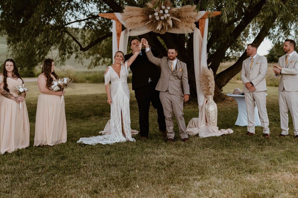 Bride and groom raise hands in celebration with wedding party under a boho arch on a sunny day.