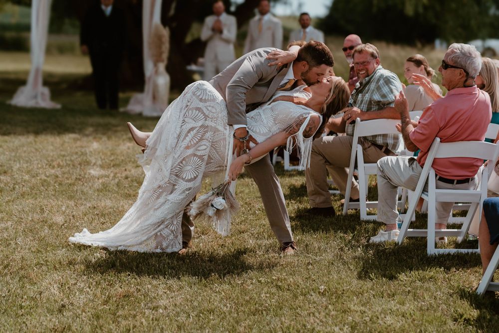 Bride and groom share a romantic dip kiss during outdoor wedding ceremony with guests seated on white chairs.