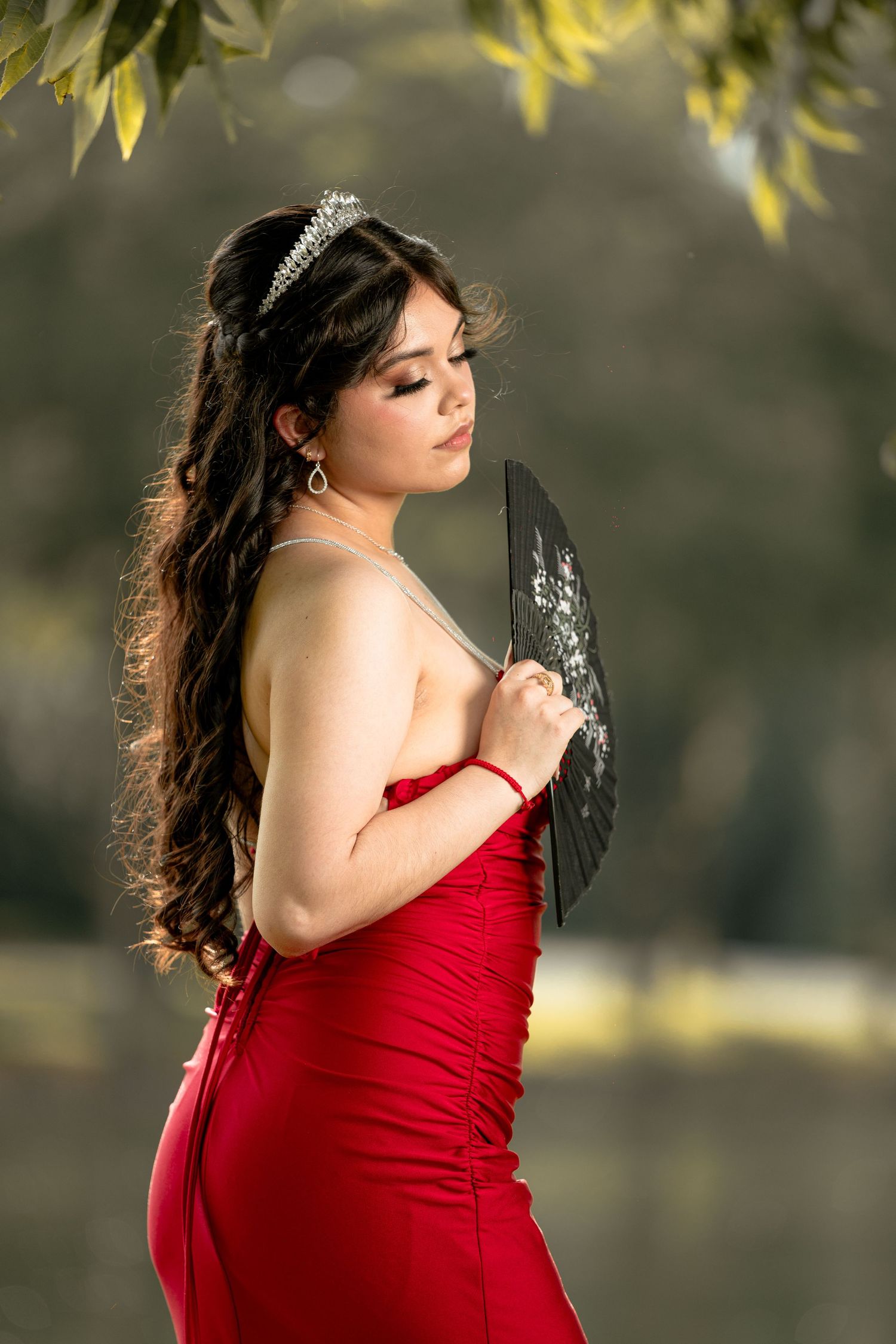 A person in a red evening gown poses elegantly with a black fan while wearing a delicate tiara against a soft bokeh background.
