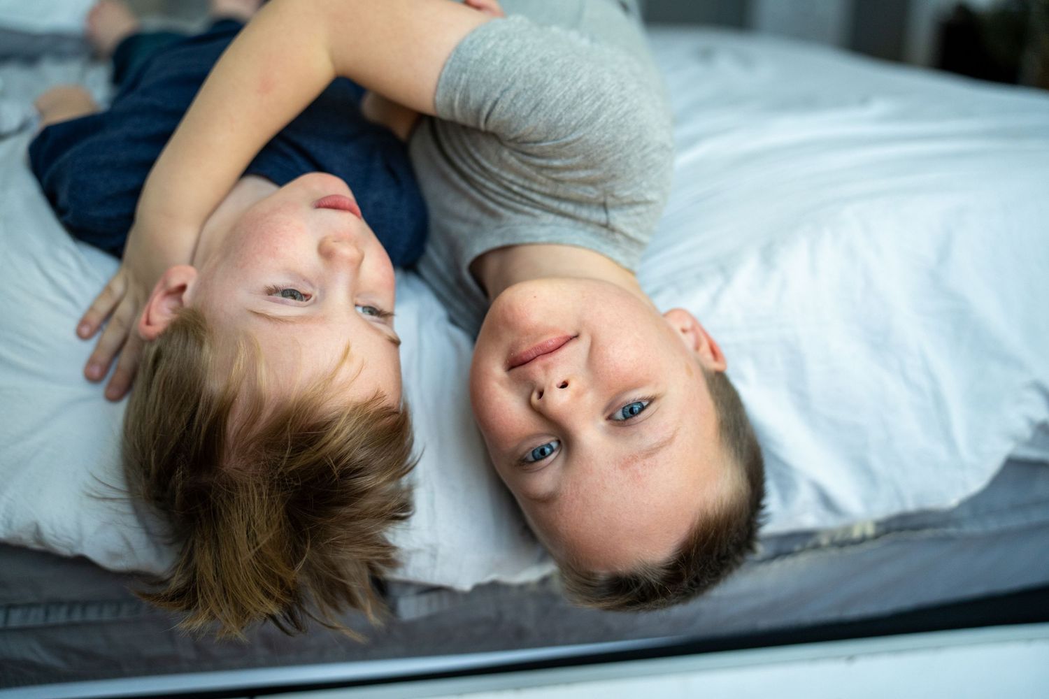 Two children lie close together on bed looking up at camera with joyful expressions while wearing casual clothes. In-home session.