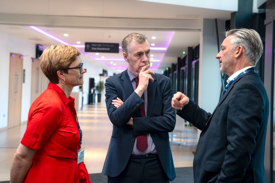 Three professionals in formal attire engaged in discussion in a modern conference center hallway.