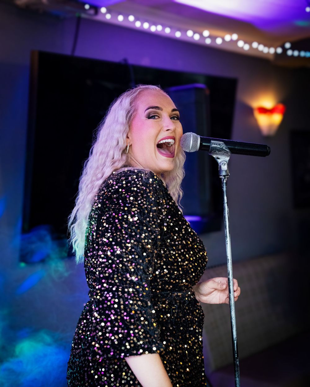 A performer in a sparkly black dress sings passionately on stage under purple and blue lighting.