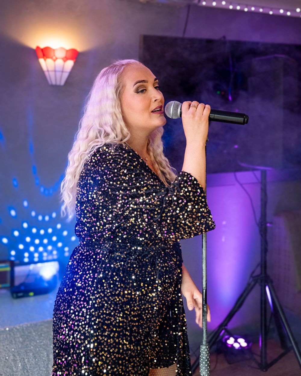 A performer in a sparkly blue sequin dress sings into a microphone on stage with purple lighting in the background.