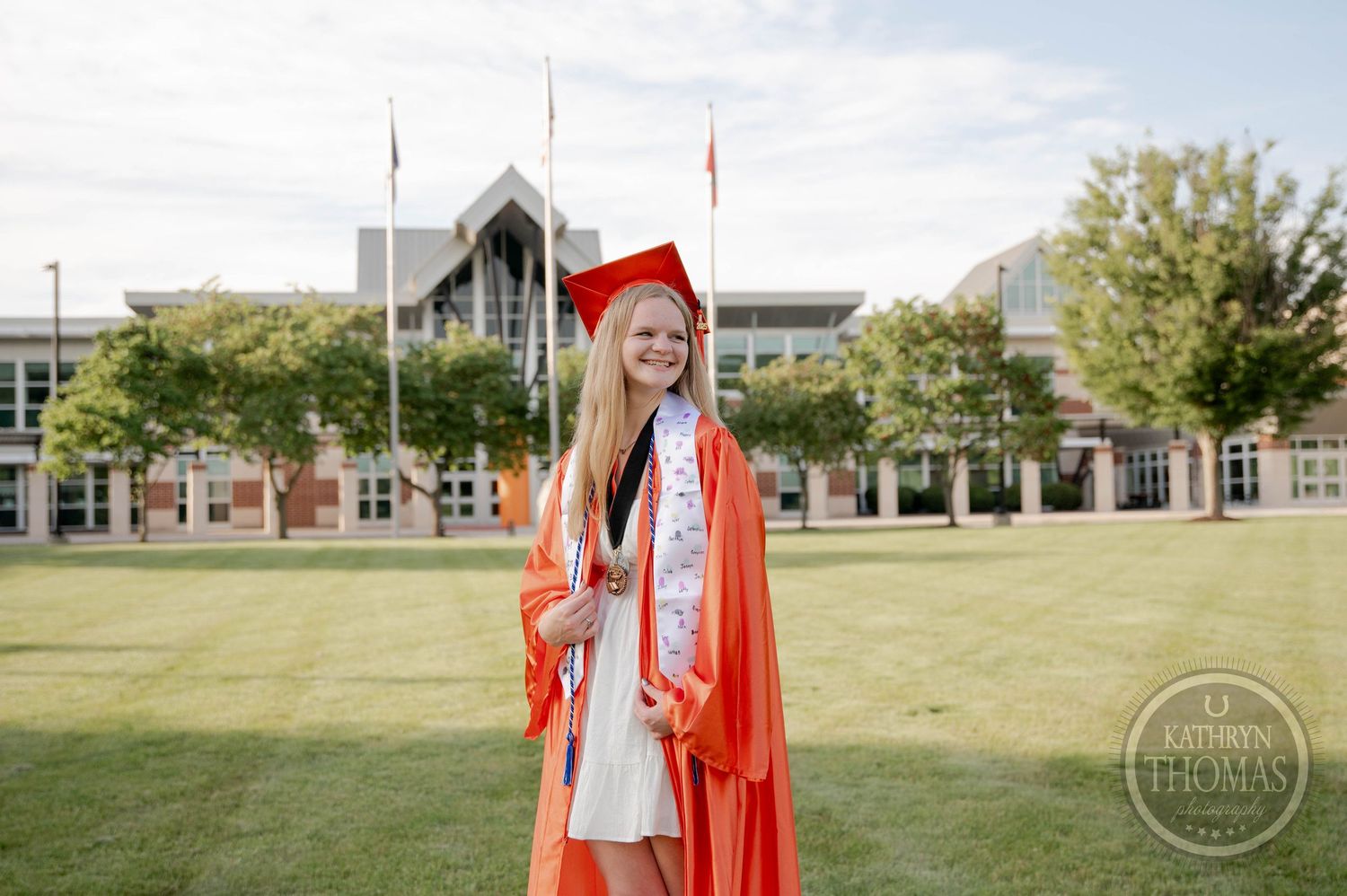 Graduate in bright orange cap and gown poses on sunny grass lawn in front of academic building during commencement.