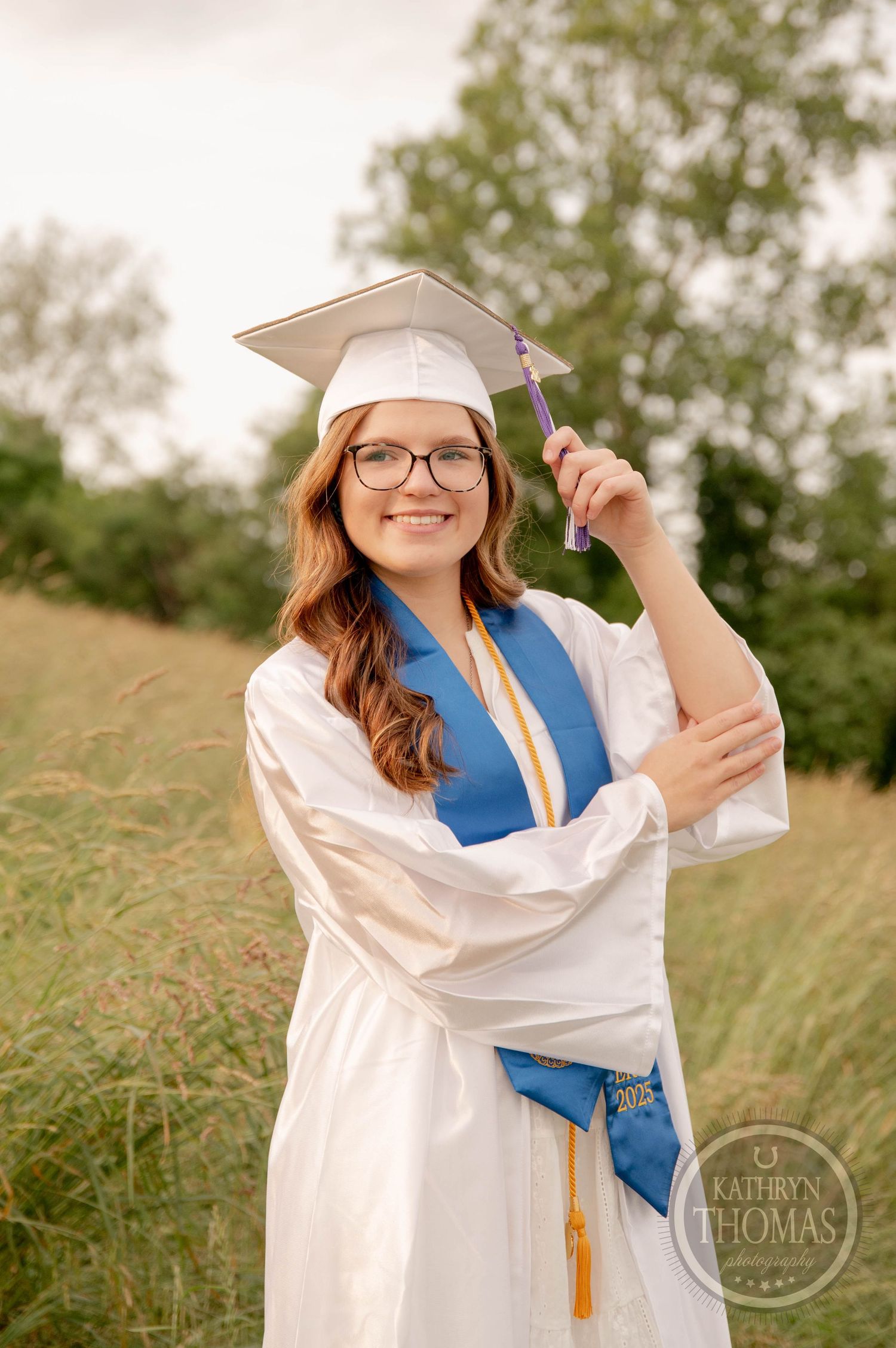 Graduate in white cap and gown with blue stole poses in grassy field during outdoor graduation photo session.