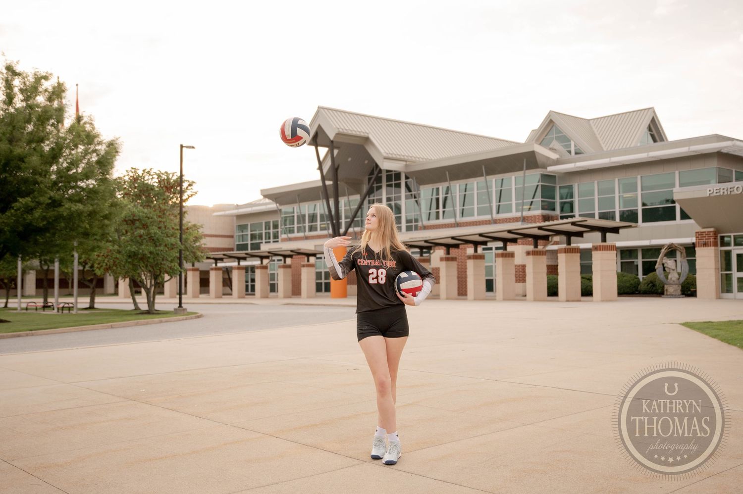 A figure in a black dress holds a basketball outside a modern campus athletic facility on a sunny day.