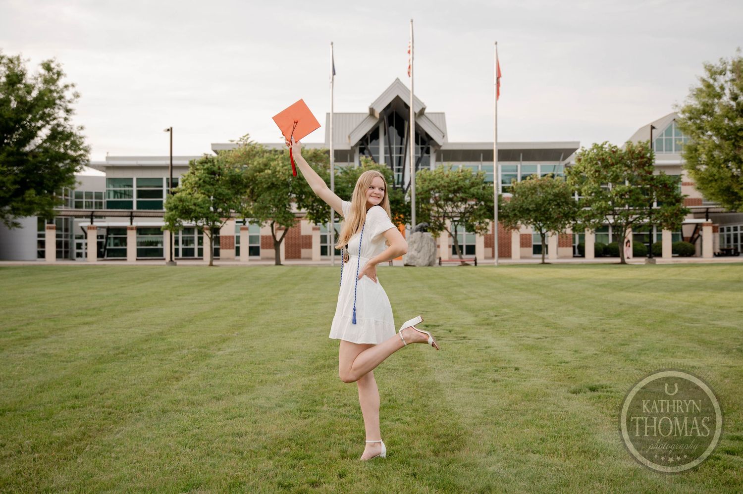 A person in a white gown twirls playfully with a bouquet on the green grass near a campus-style building.