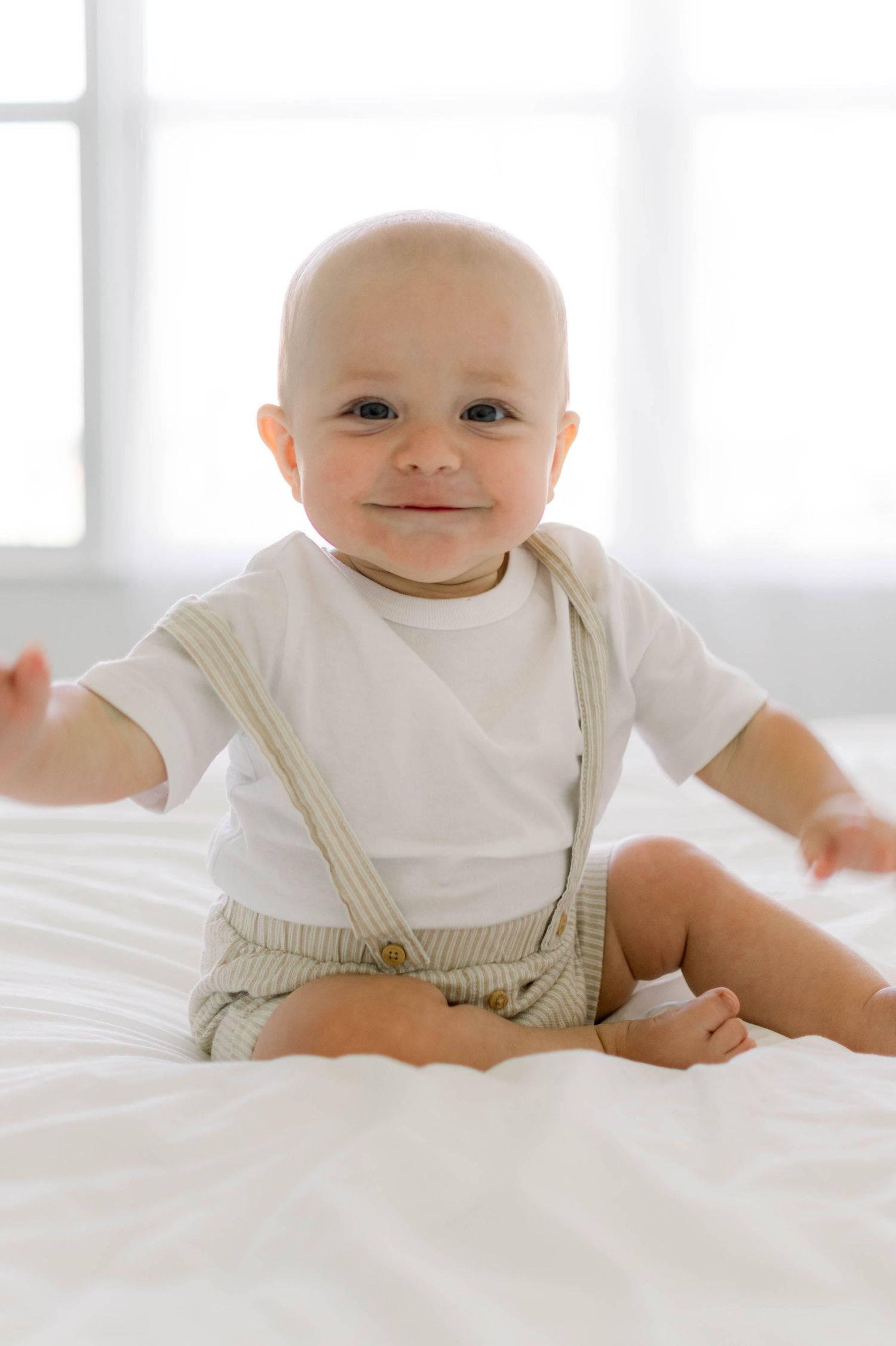 Sweet baby wearing white t-shirt and beige suspender shorts sits smiling on white bedding in bright room.