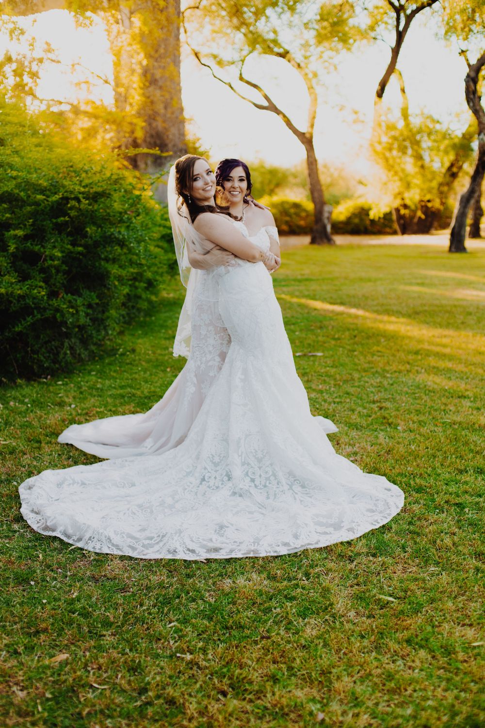 Two people embrace while wearing elegant white wedding gowns on a sunlit lawn with golden autumn trees in the background.