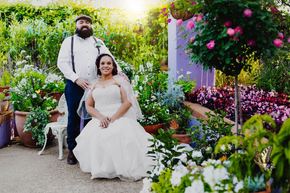 A wedding couple sits in a lush garden setting surrounded by colorful flowers and greenery with sunlight peeking through.