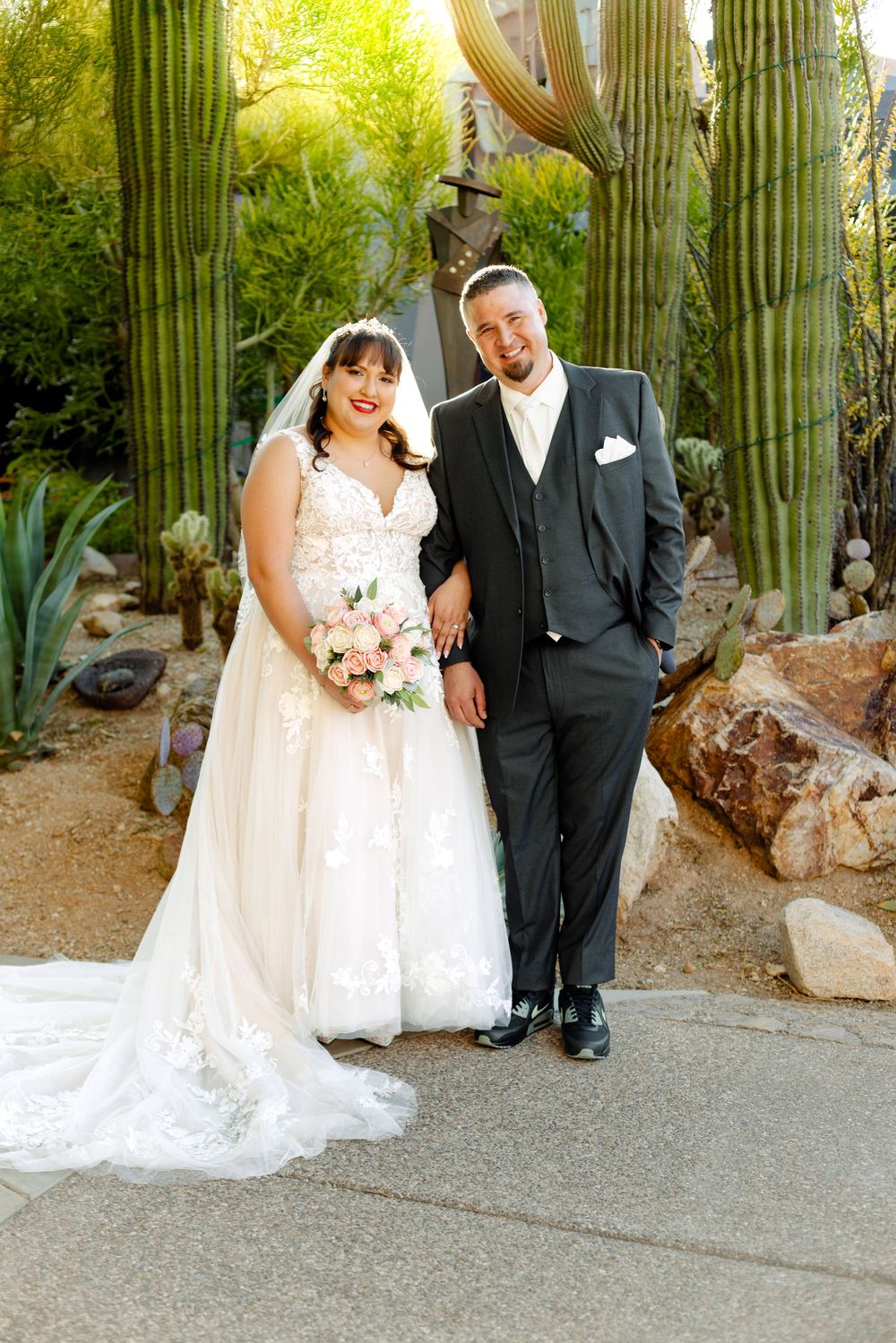 Wedding couple poses by towering saguaro cacti and desert landscaping in an outdoor garden setting.