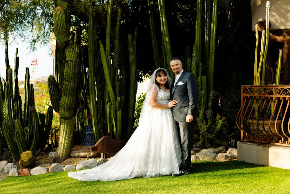 A couple in wedding attire poses together in front of tall green cactus plants and trees in a garden setting.