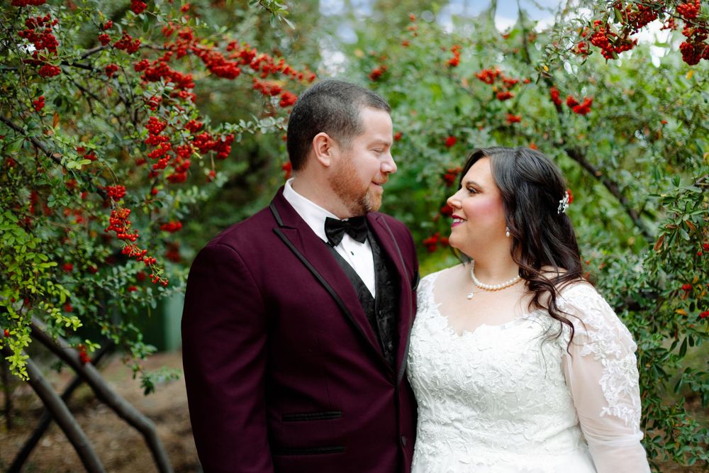 A romantic wedding couple stands together among vibrant red berry bushes in an outdoor garden setting.