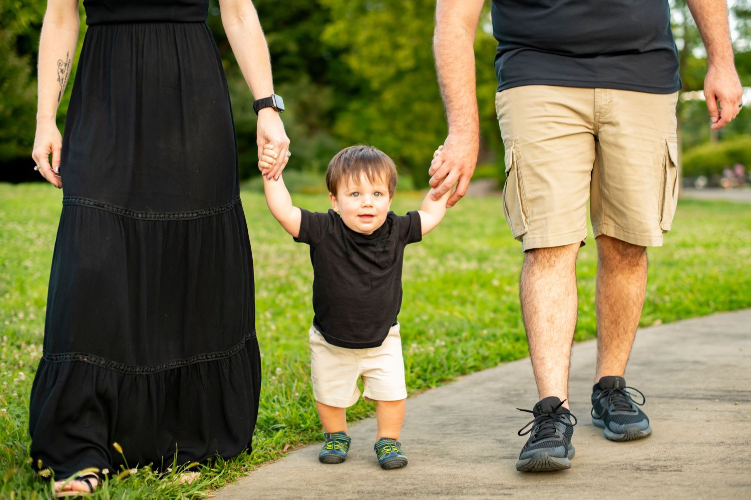 A child wearing a black shirt and khaki shorts stands between their parents on a paved path in a park.