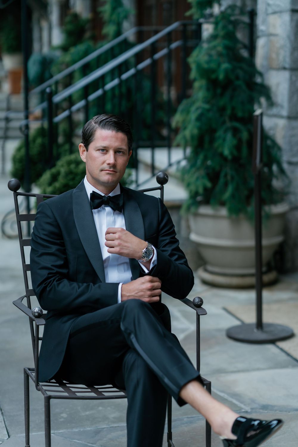 A person in a black tuxedo and bowtie sits casually on a dark metal chair outdoors. old edwards inn north carolina