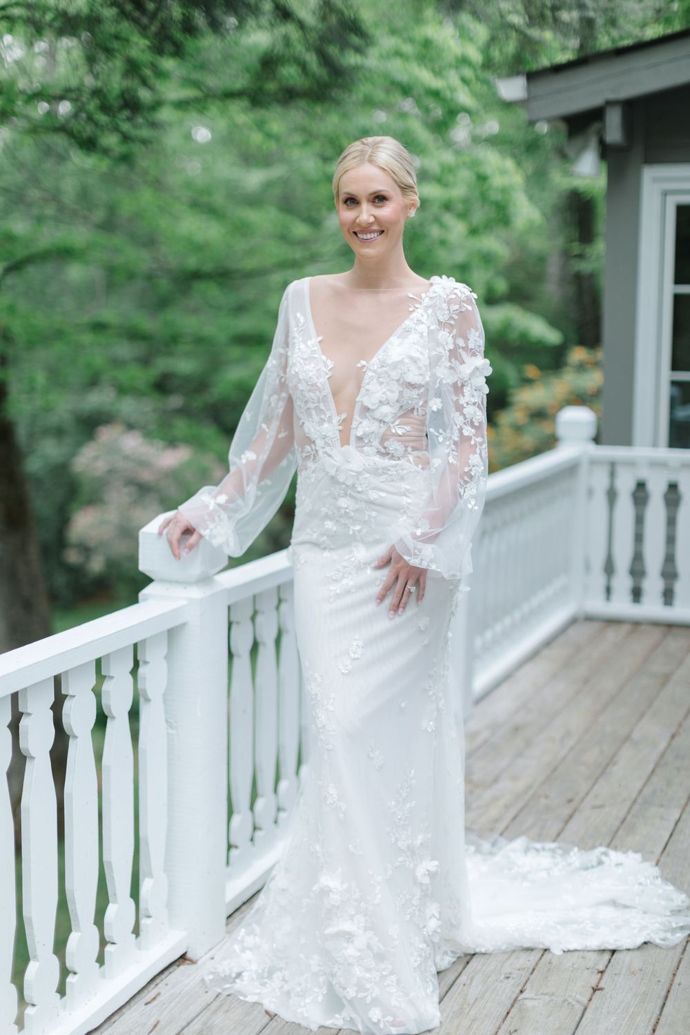 A bride in a long-sleeved lace wedding gown poses gracefully on a white porch railing surrounded by greenery. Old Edwards Inn