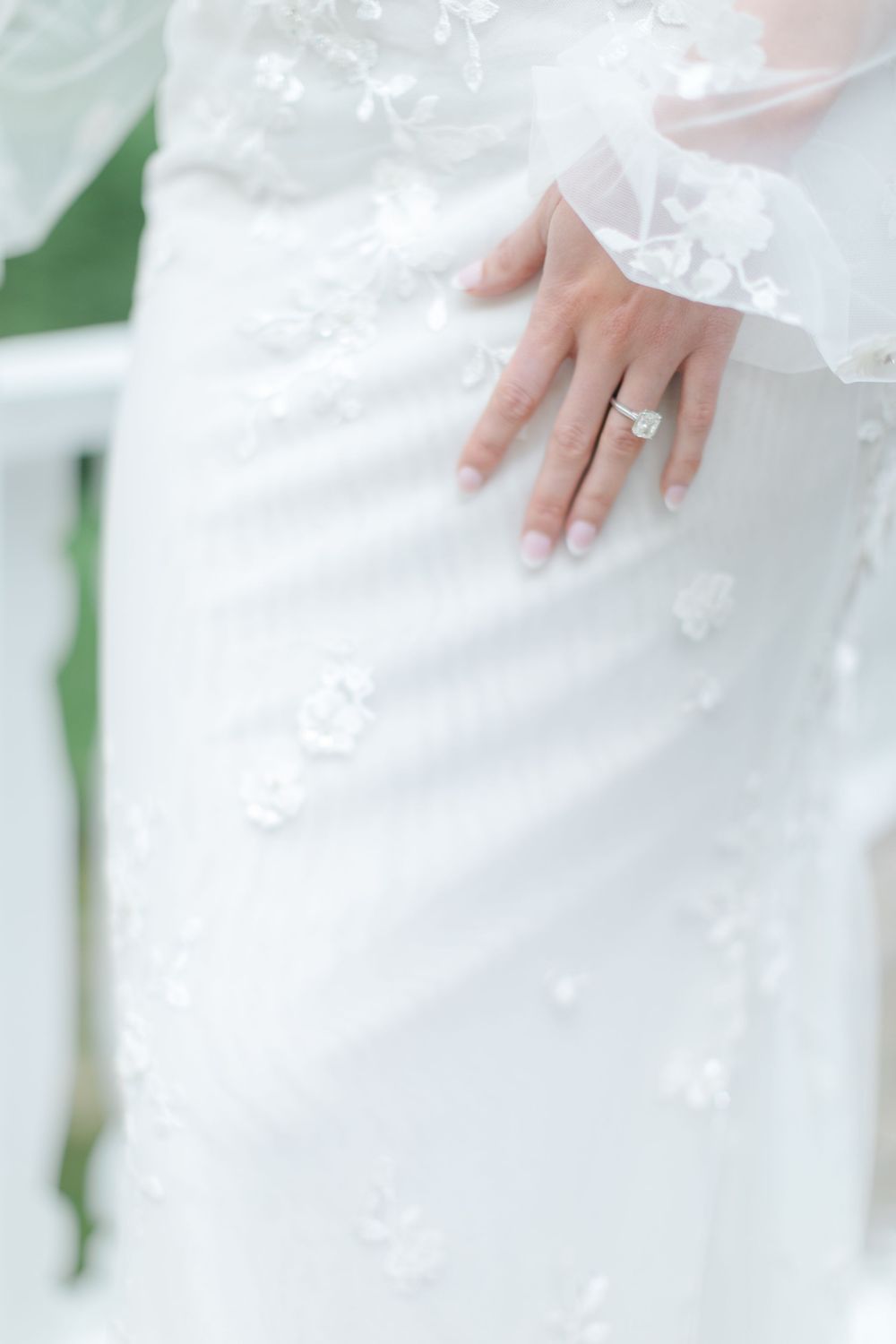 Close-up detail of a white lace wedding dress with delicate floral patterns and a hand wearing an engagement ring.