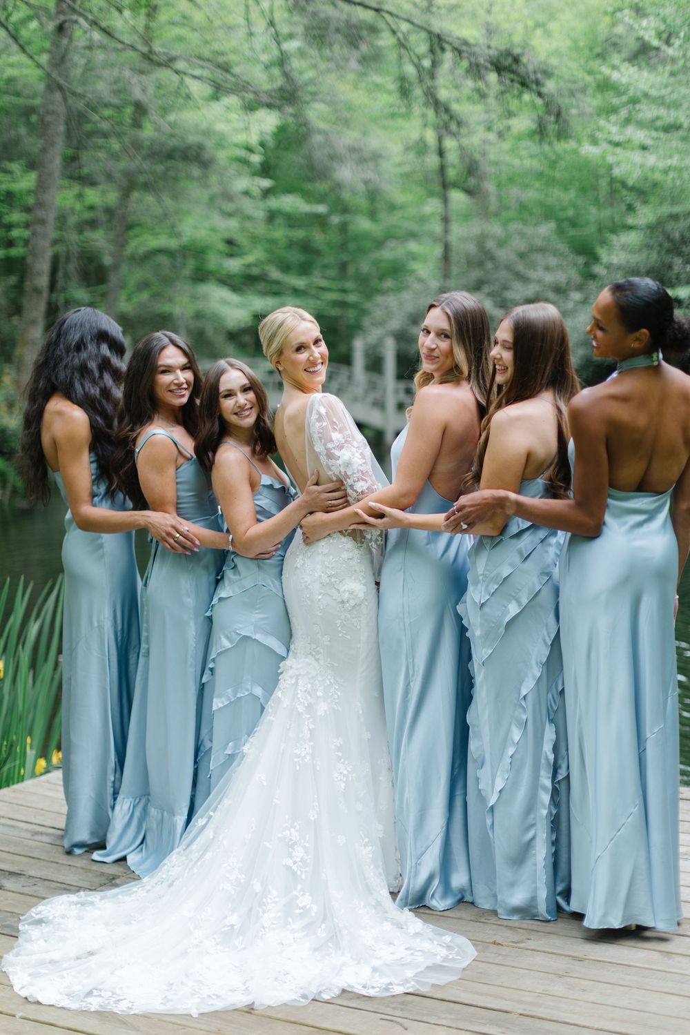 A bridal party in light blue bridesmaid dresses and white wedding gown pose together on a wooden deck surrounded by trees. Old Edwards Inn