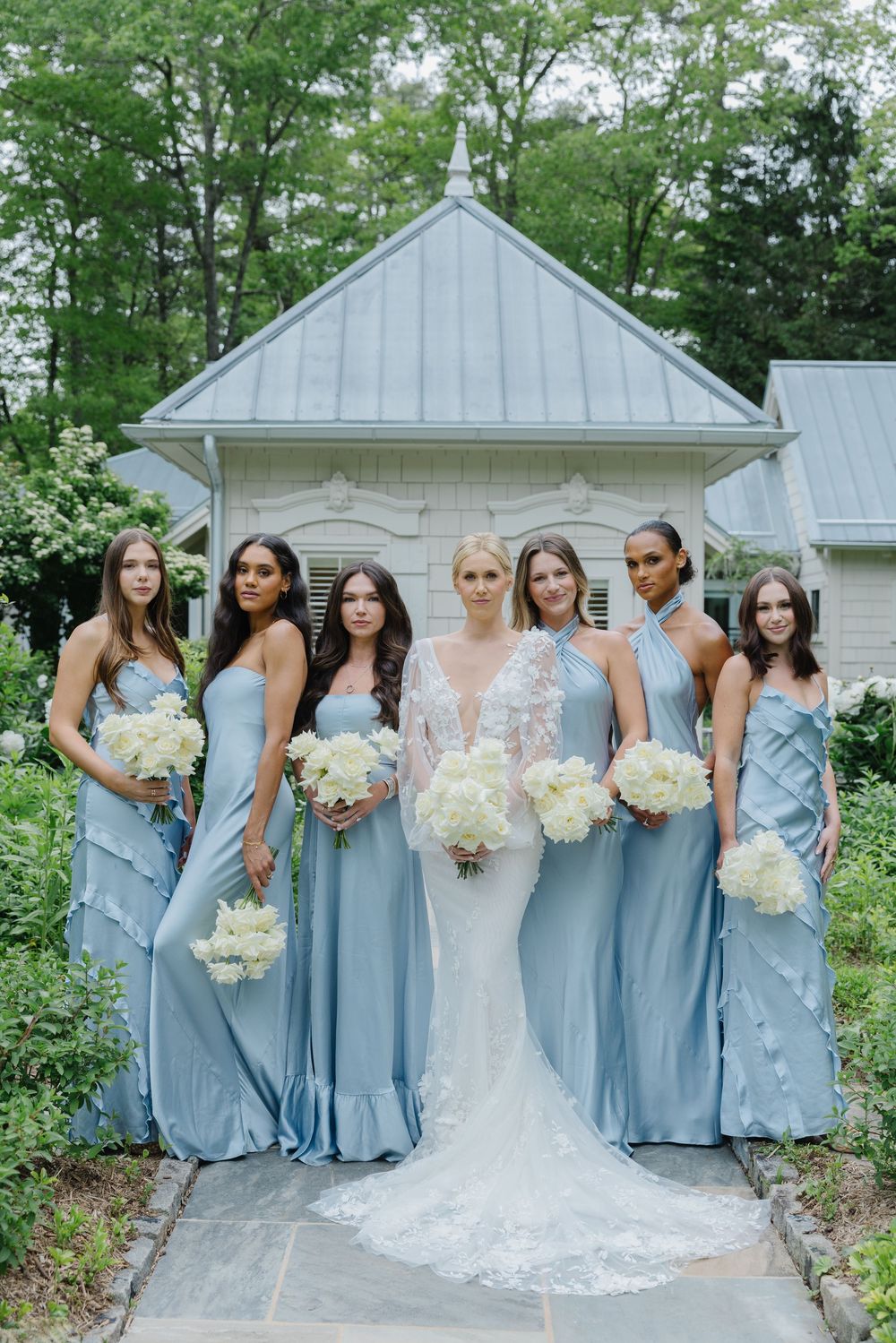Wedding party in light blue bridesmaid dresses and white bridal gown pose in front of white chapel building and greenery. old edwards inn
