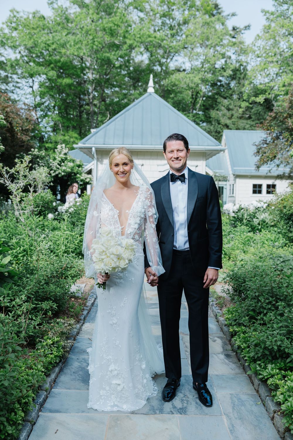 A wedding couple stands on a garden pathway surrounded by lush green shrubbery and white chapel buildings. old edwards inn