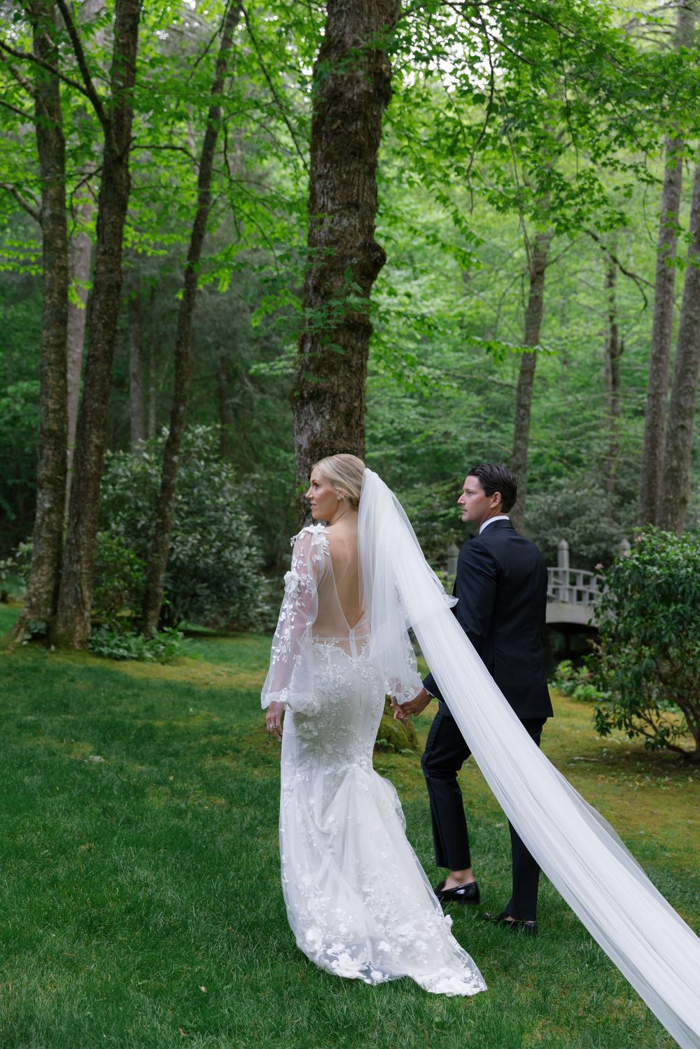 A couple stands on a curved wooden footbridge spanning a tranquil pond surrounded by lush green. bride and groom old edwards inn