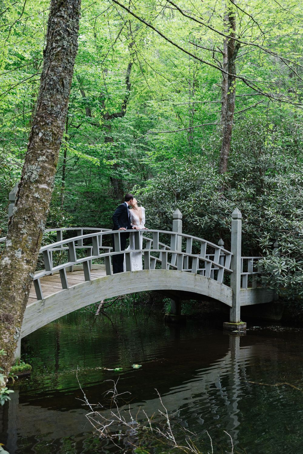 A couple stands on a curved wooden footbridge spanning a tranquil pond surrounded by lush green. bride and groom old edwards inn