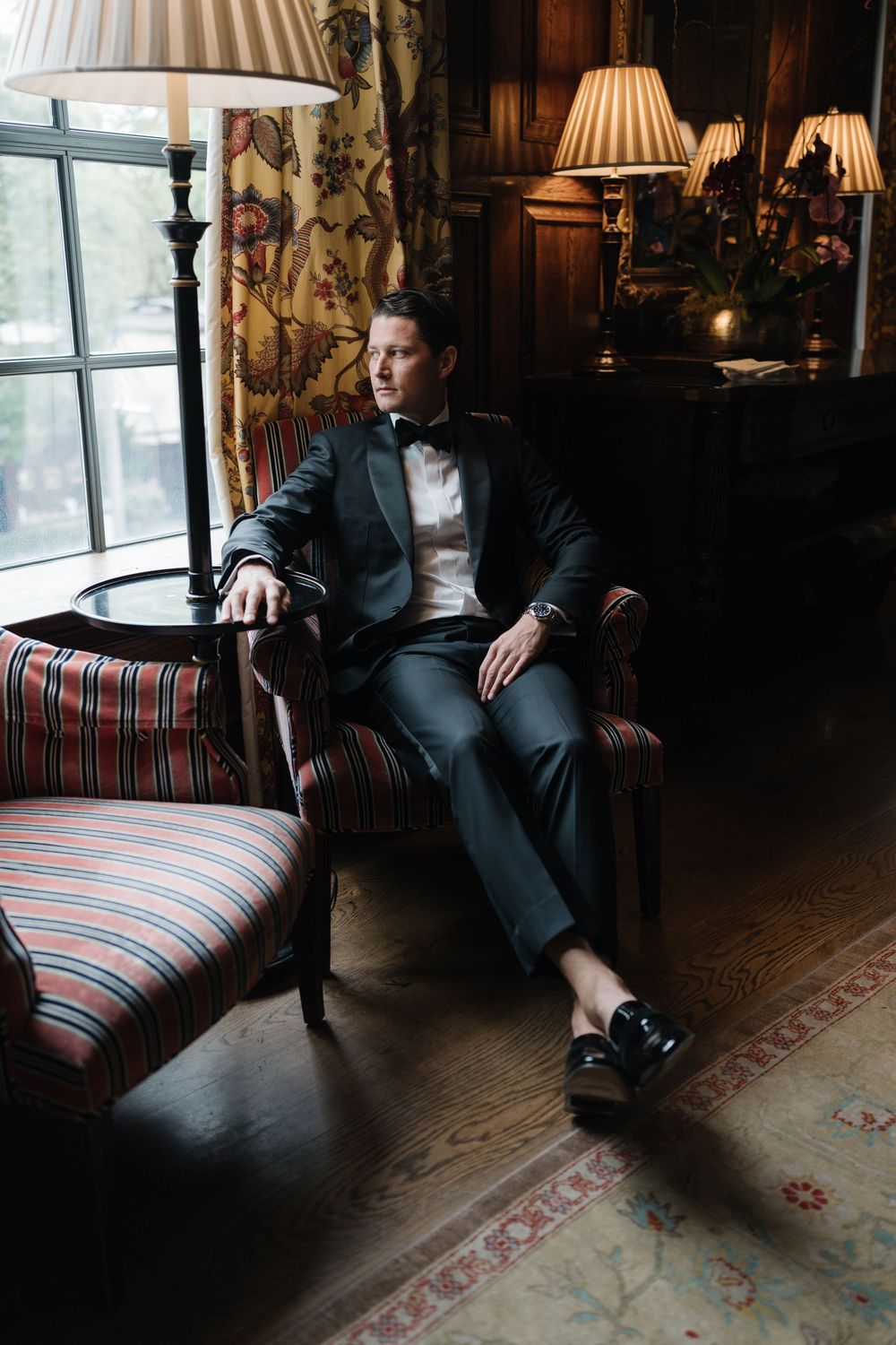 Moody interior photo of someone in a dark suit sitting in a vintage armchair near a window with striped upholstery and floral curtains.