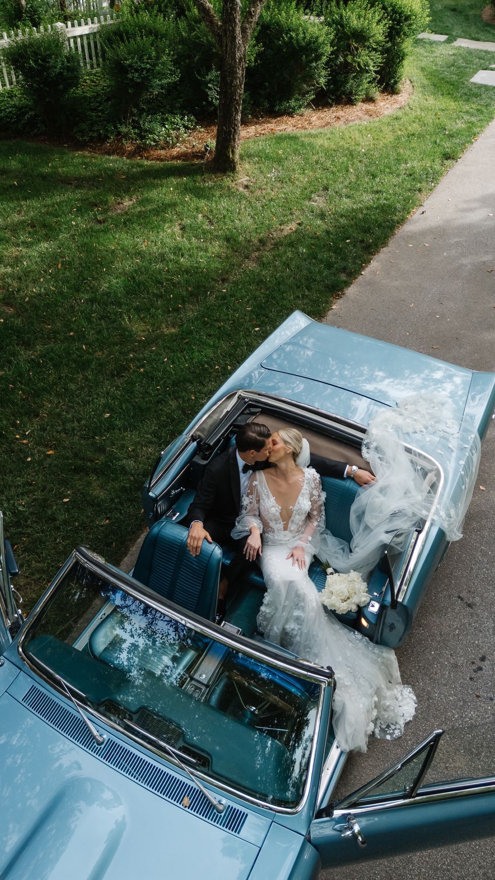 A couple in a classic light blue convertible car is photographed from above during what appears to be a wedding celebration.