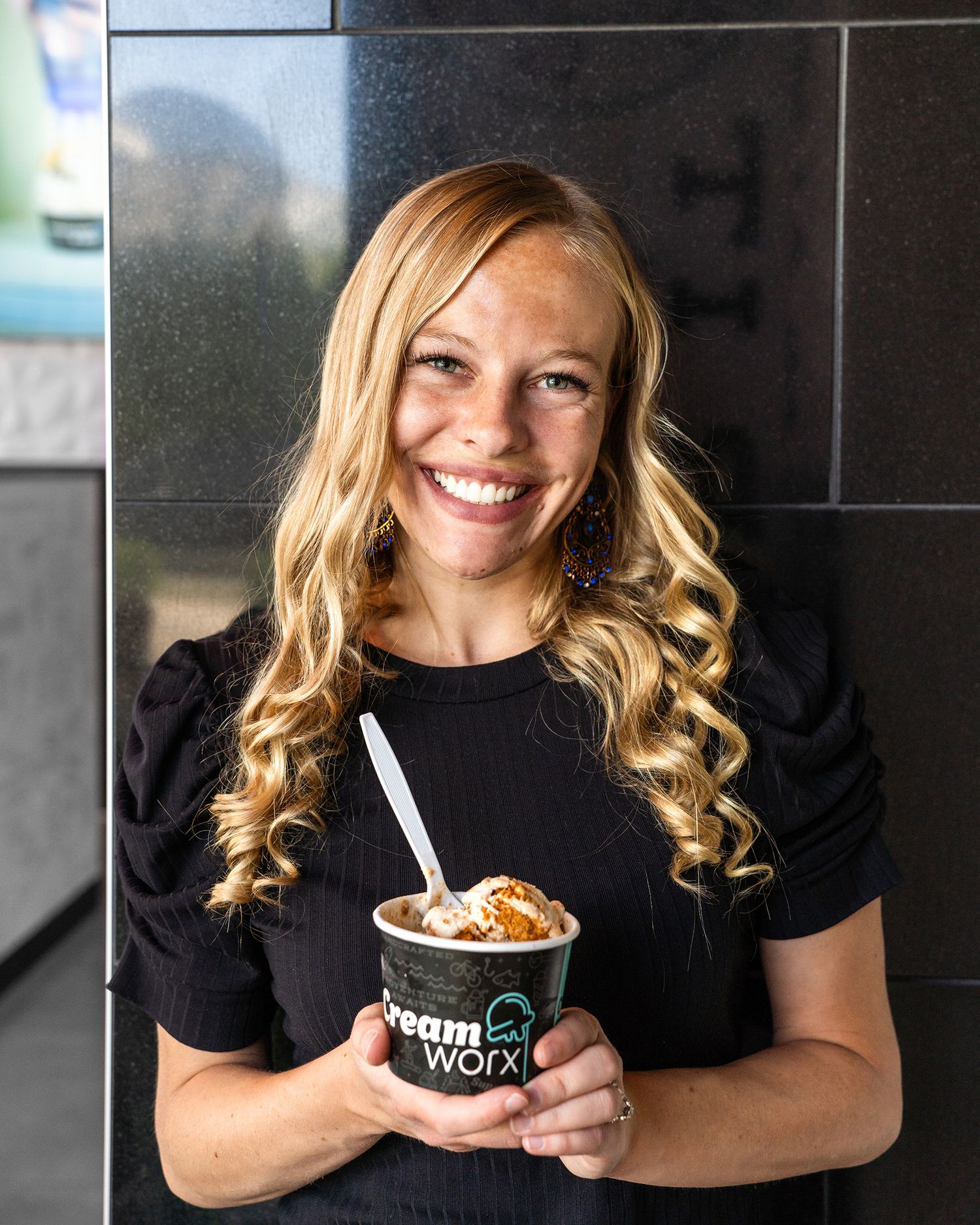 A person with blonde wavy hair wearing a black shirt smiles while holding a branded ice cream cup and spoon.