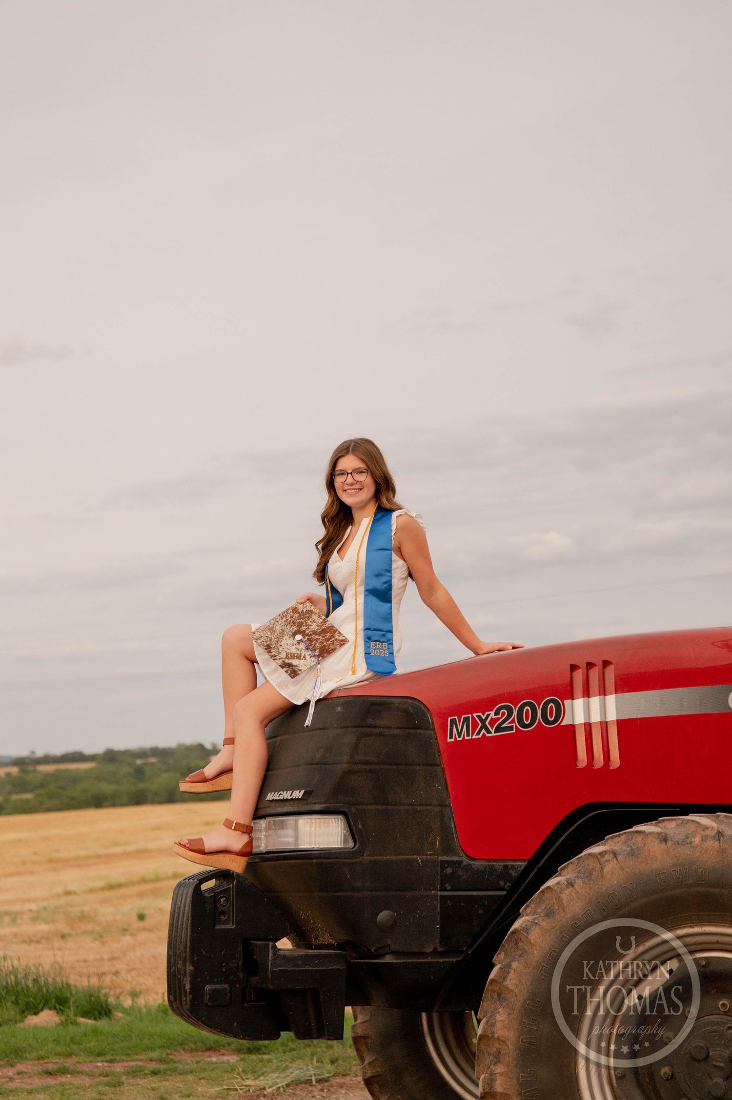 A person sits on a red Case IH MX200 tractor in a rural field setting with their cat on a cloudy day.