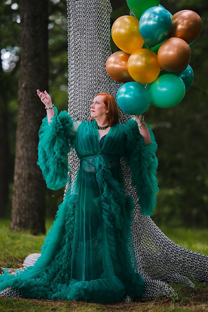 A figure in a flowing emerald green tulle gown poses dramatically with copper and turquoise balloons in a forest setting.