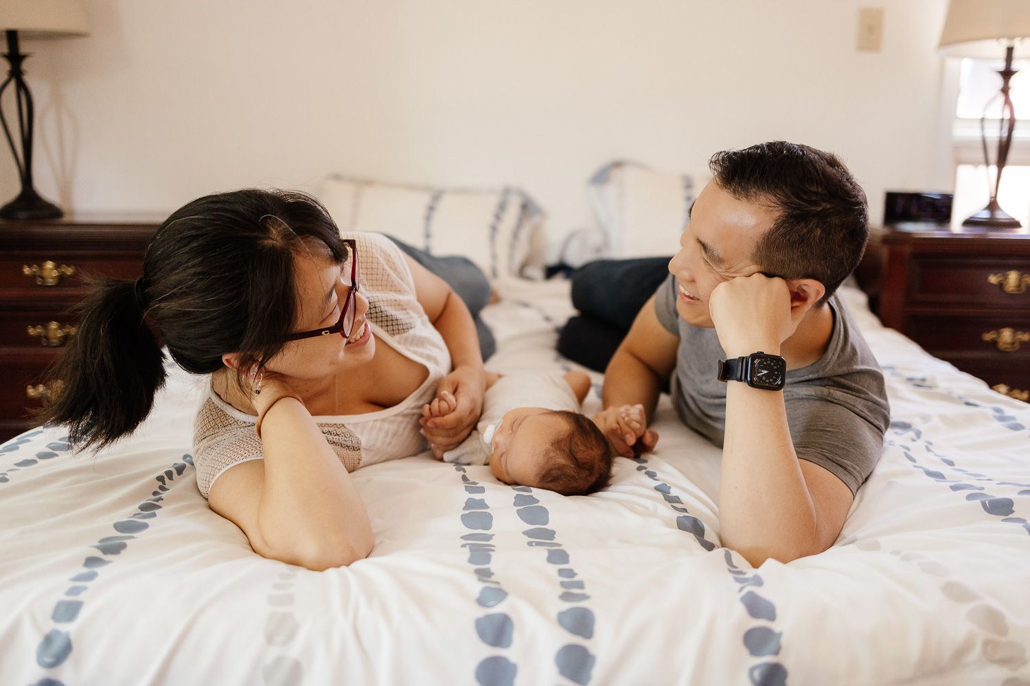 Parents smile at each other while laying on their bed with their baby in between them