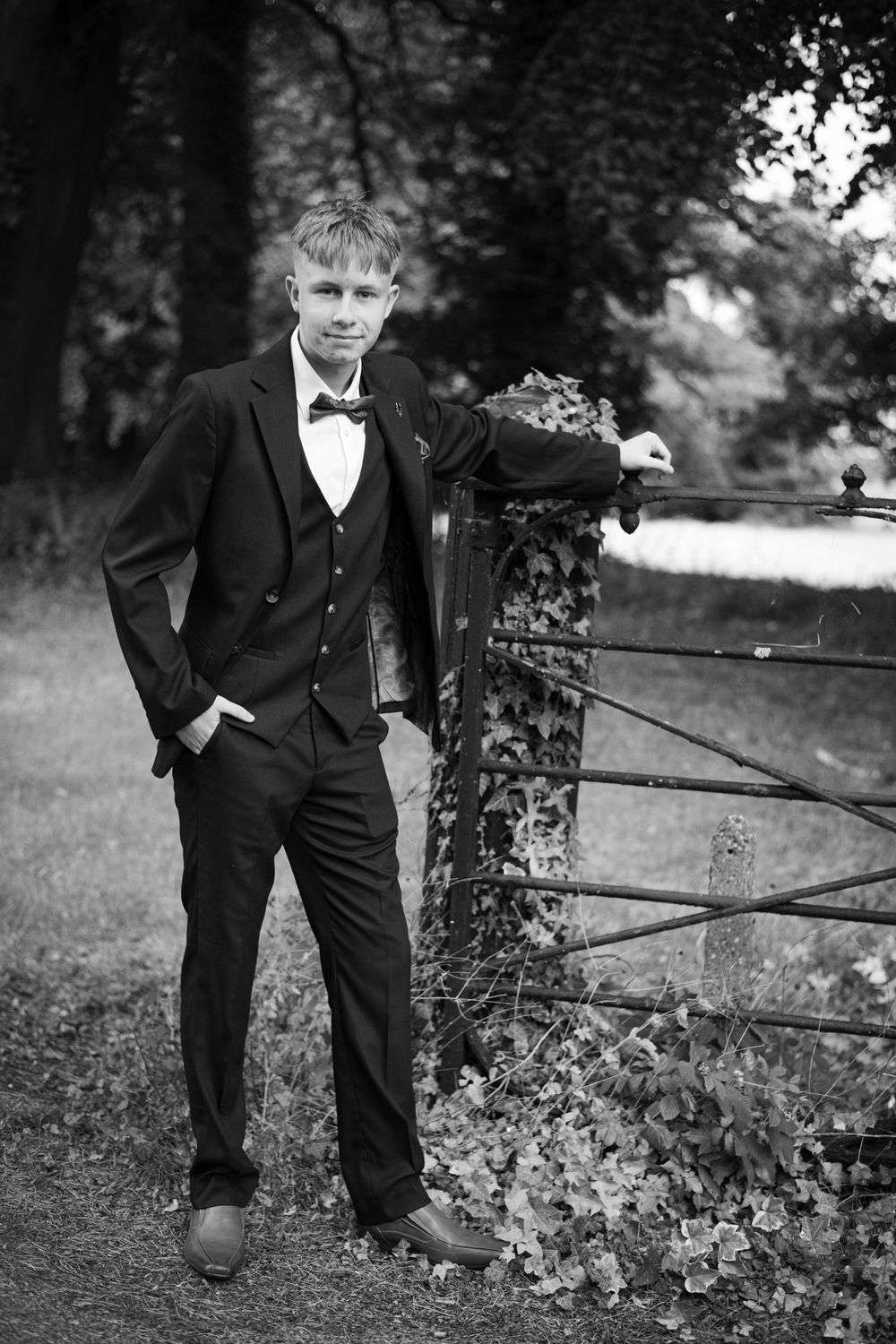 A black and white formal portrait of a young person in a tuxedo leaning against a rustic gate outdoors.