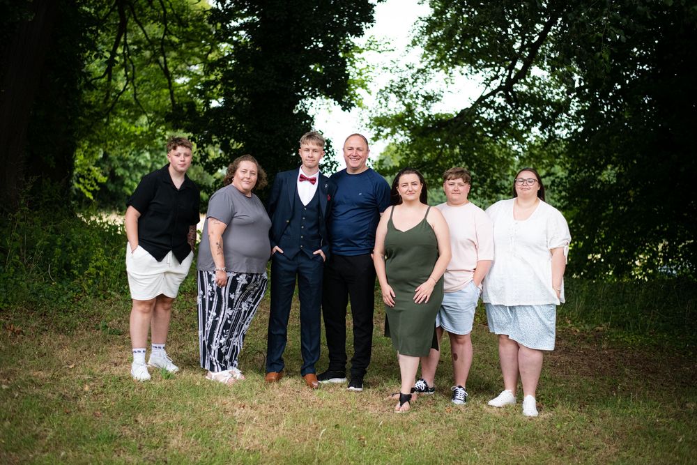 A group of people pose together outdoors on grass with green trees in the background on a sunny day.